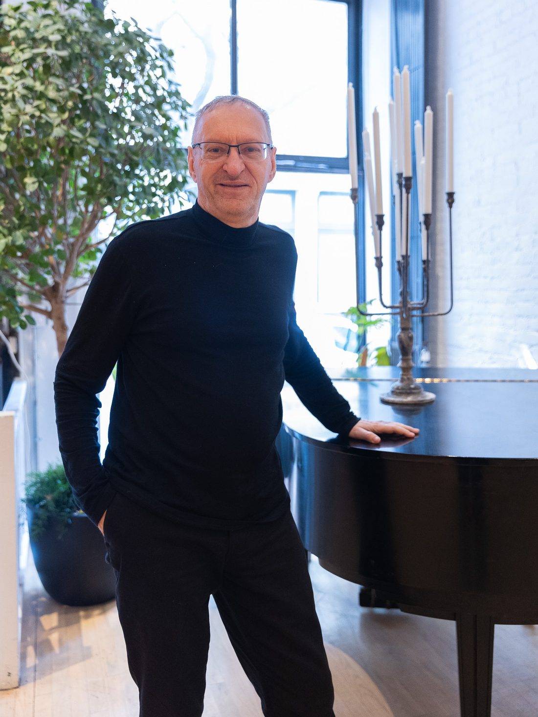 Pierre Boutquin standing beside a grand piano in a bright loft, one hand resting on the lid
