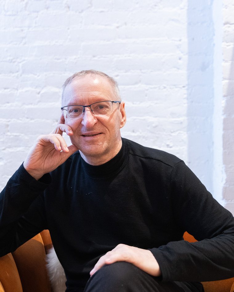 Pierre Boutquin seated, hand resting against his temple, in a bright loft with a white brick wall